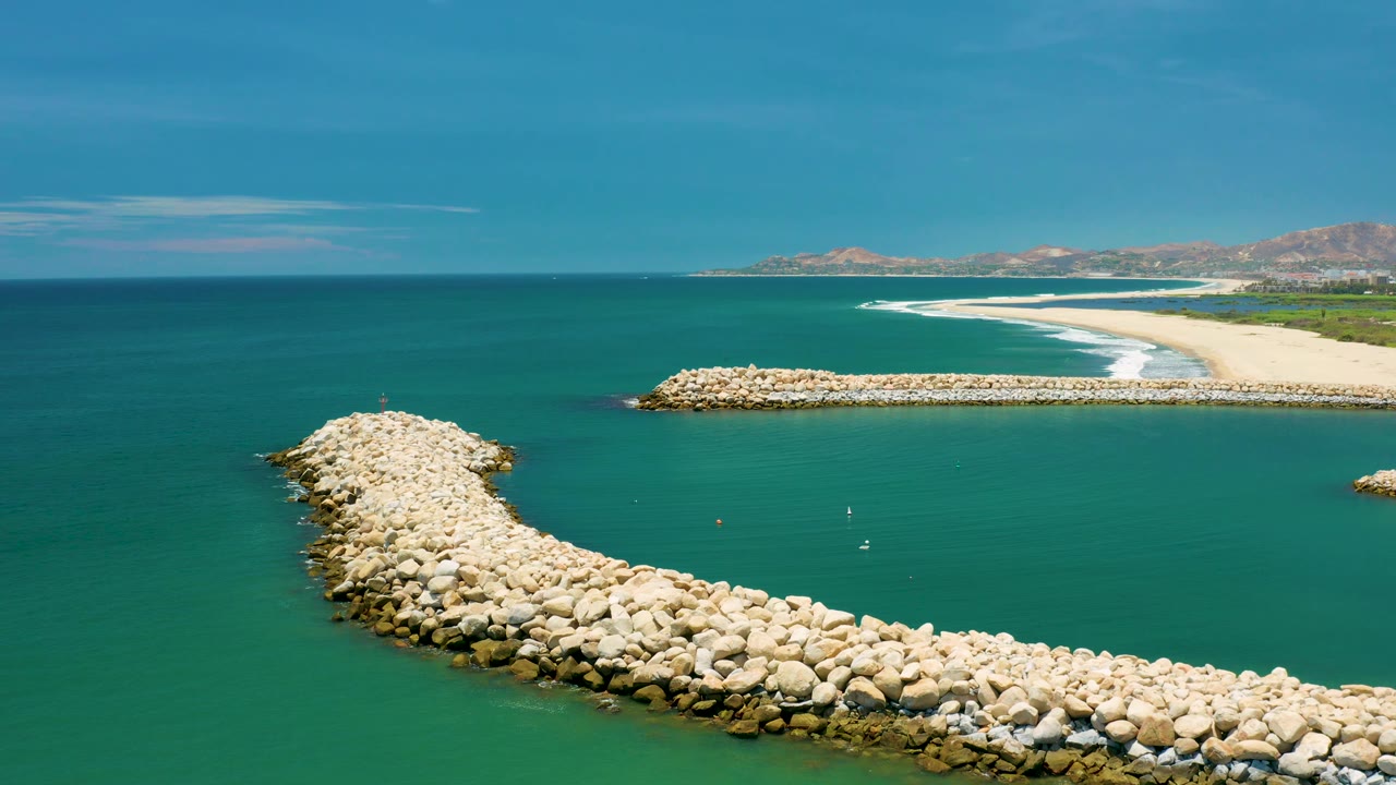 Aerial view in San Jose Del Cabo, Mexico, with the rocky breakwater spanning the lower frame, calm green water inside the curve, white surf on the beach beyond, and no visible people.