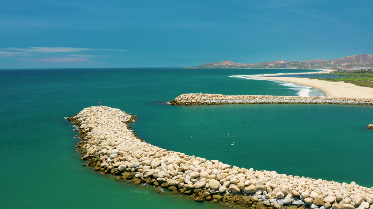 Mid-clip aerial frame in San Jose Del Cabo, Mexico, showing a boulder arm extending from the lower edge into the ocean, small white markers on the water, beach surf, hills, and no visible people.