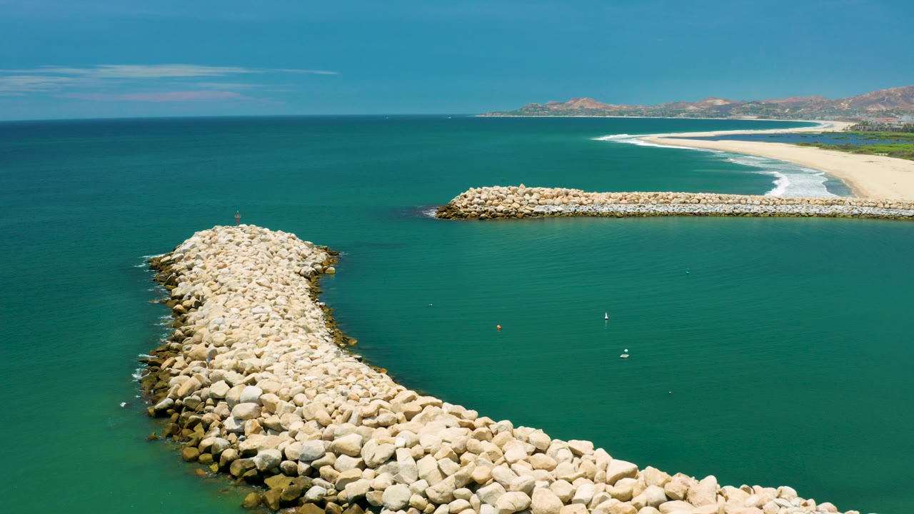 Aerial frame over San Jose Del Cabo with a curved white boulder breakwater in the foreground, a straight rock barrier near the beach, blue-green water, distant hills, and no visible people.