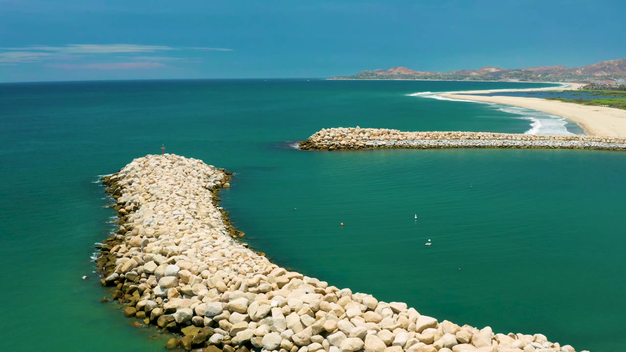 Drone view of white stone jetty extending into calm blue-green water near a beach.