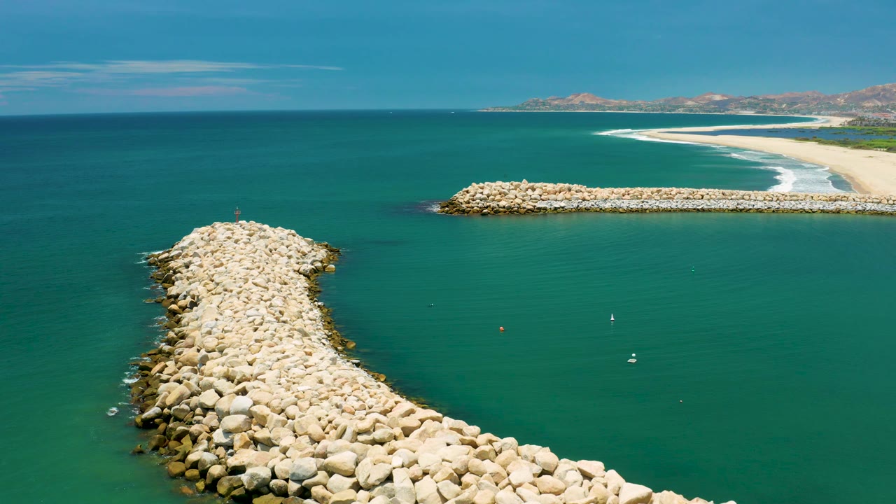 A high coastal view in San Jose Del Cabo shows the curved boulder jetty lower left, the straight breakwater near the surf line, turquoise water across the frame, low hills on the horizon, and no visible people.