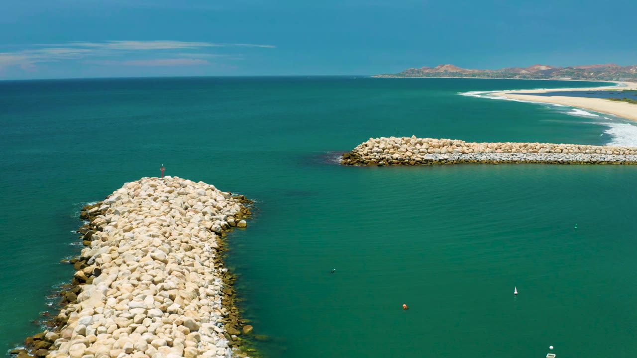 Aerial frame from San Jose Del Cabo, Mexico, with the main white rock jetty centered lower in the view, a horizontal breakwater to the right, turquoise-green water, far beach, and no visible people.