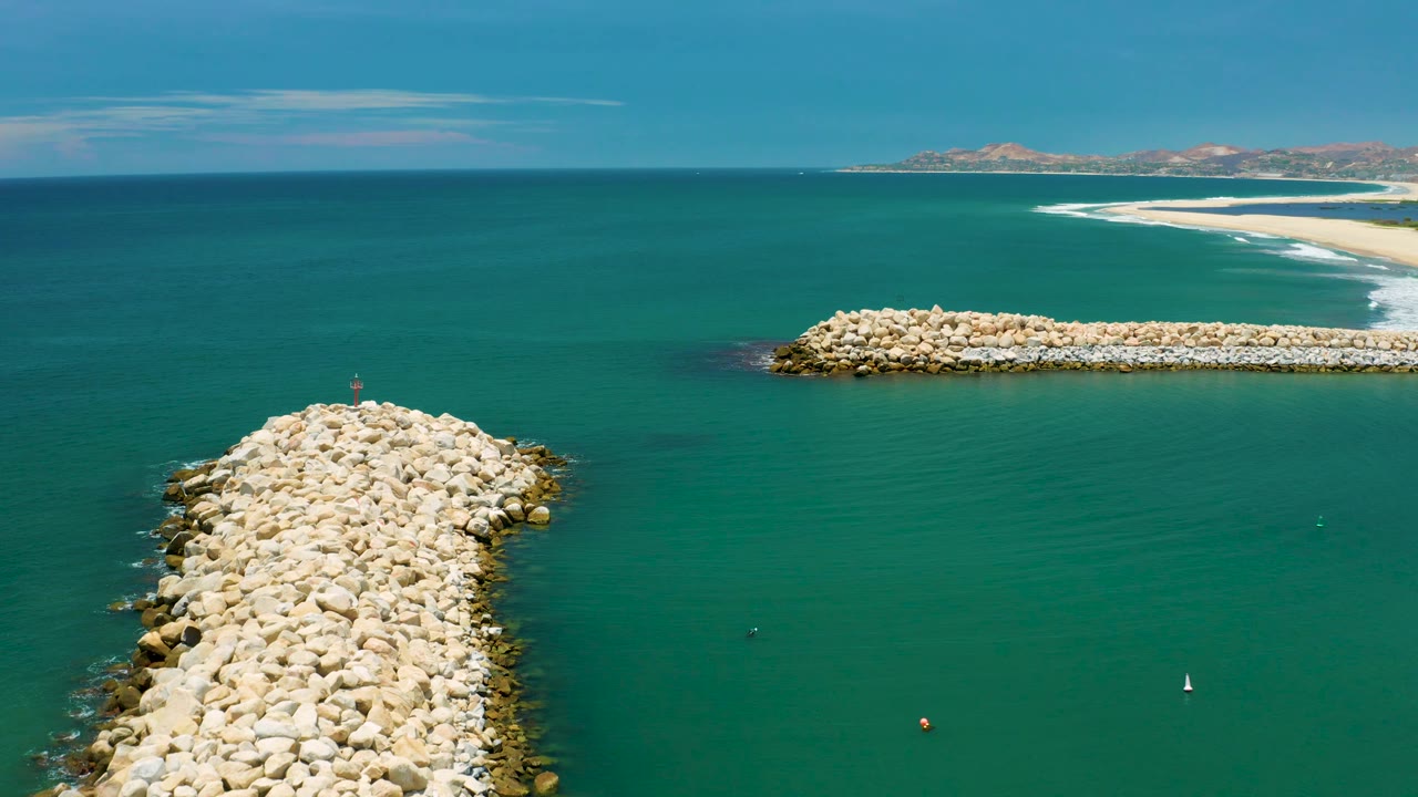 Aerial view over San Jose Del Cabo, Mexico, with the foreground white rock jetty slightly left of center, a long breakwater in the upper right, a pale surf line on the distant beach, and no visible people.