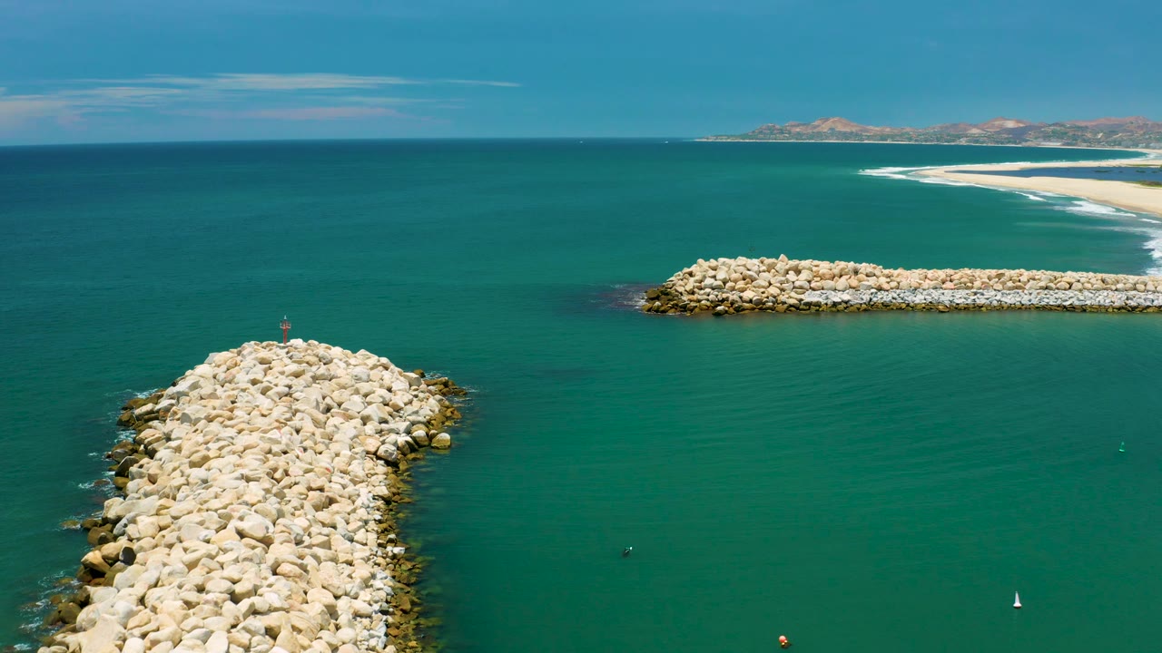 San Jose Del Cabo, Mexico aerial frame with the main white boulder jetty entering from the lower left, calm green water across the center, a right-side breakwater, distant sand, and no visible people.