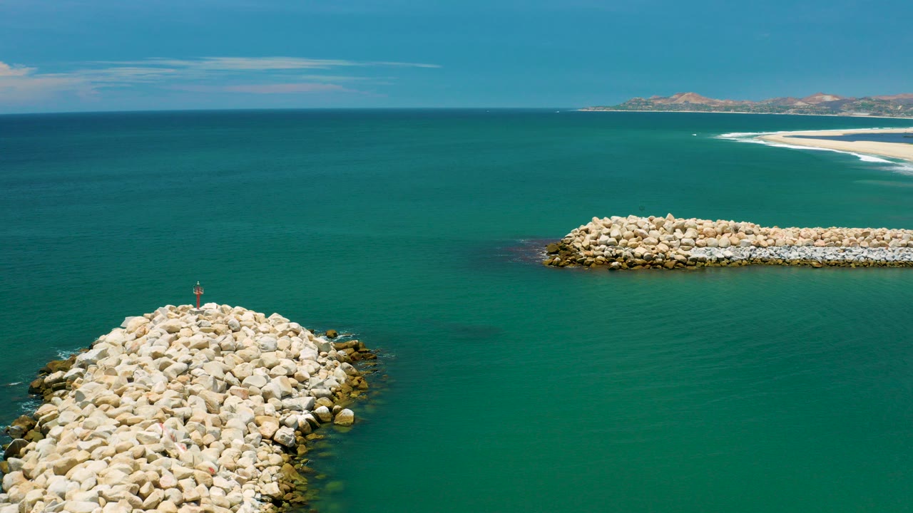Aerial frame from San Jose Del Cabo, Mexico showing turquoise water, a foreground rock breakwater at lower left, a second rock jetty across the middle, distant shore, and no visible people.