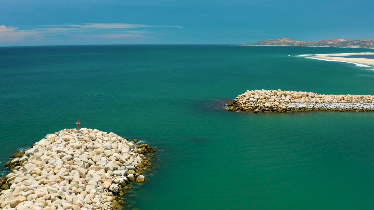 Aerial frame from San Jose Del Cabo, Mexico showing a cropped foreground rock breakwater at bottom center, a second pale rock jetty at upper right, a bright sandbar near the horizon, and no visible people.
