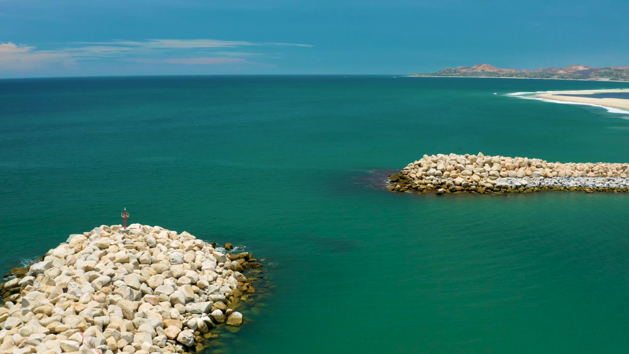 Aerial frame from San Jose Del Cabo, Mexico with foreground rocks entering from the lower left, a long pale rock jetty across the midground, open blue-green water, distant land, and no visible people.