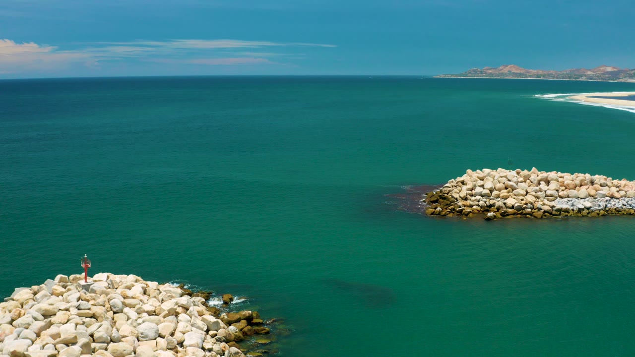 Aerial view over blue-green sea near San Jose del Cabo, Mexico, with a pale rock breakwater at the lower left, another rocky outcrop midframe, distant sand and hills on the horizon, and no visible people.