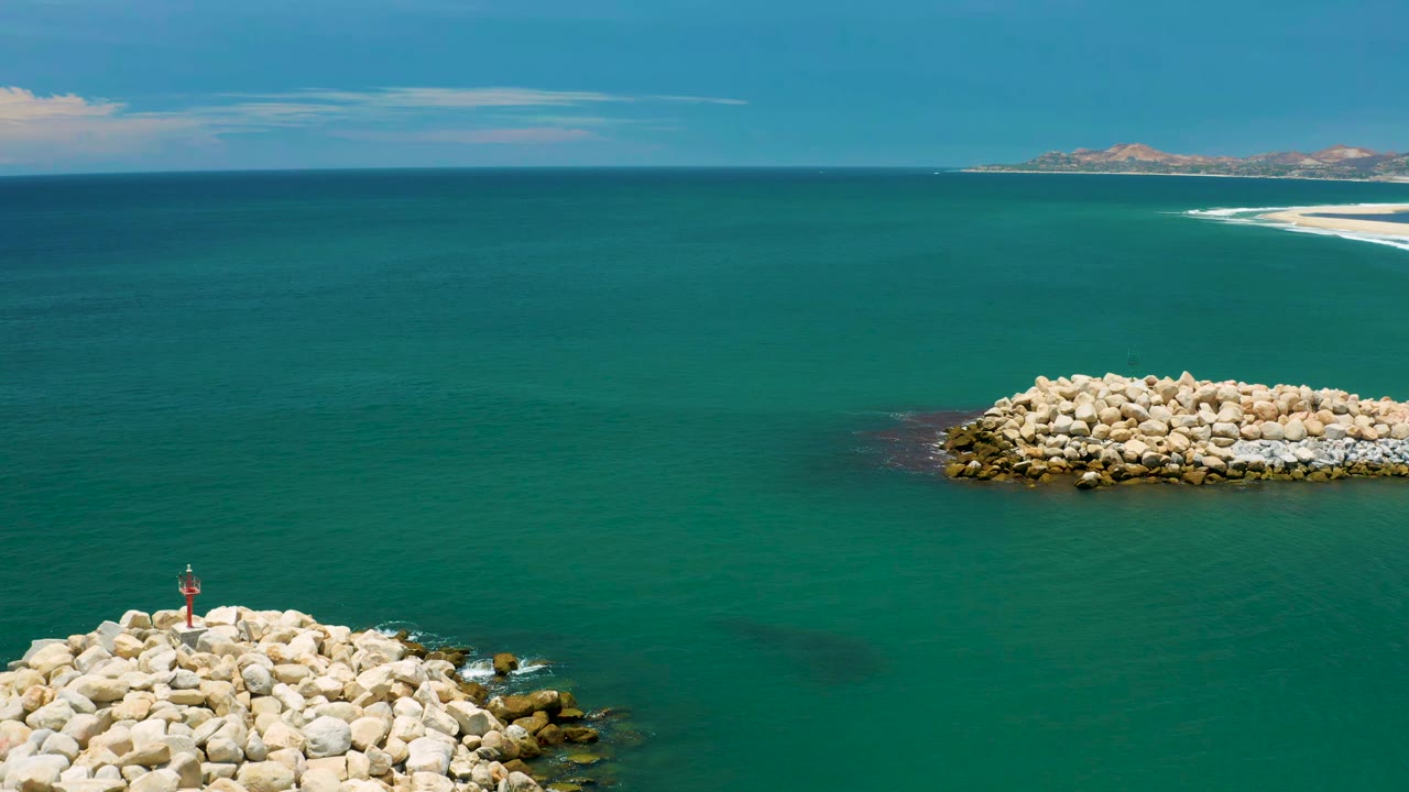 Aerial frame over San Jose del Cabo water with the foreground rocks slightly lower in view, a pale rock island across the center, distant surf along a sandy edge, and no visible people.
