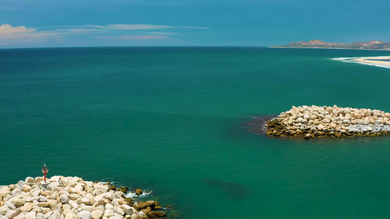 Aerial view near San Jose del Cabo with a small red marker on foreground rocks, a separate rock mound to the right, wide blue-green sea, distant coastline, and no visible people.