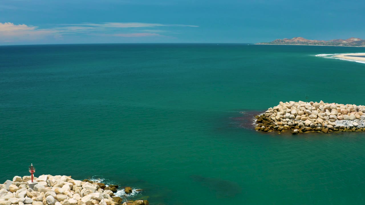 Aerial frame above San Jose del Cabo water with the foreground rocks clipped at the bottom edge, a pale rock mound right of center, distant sand and low hills, and no visible people.