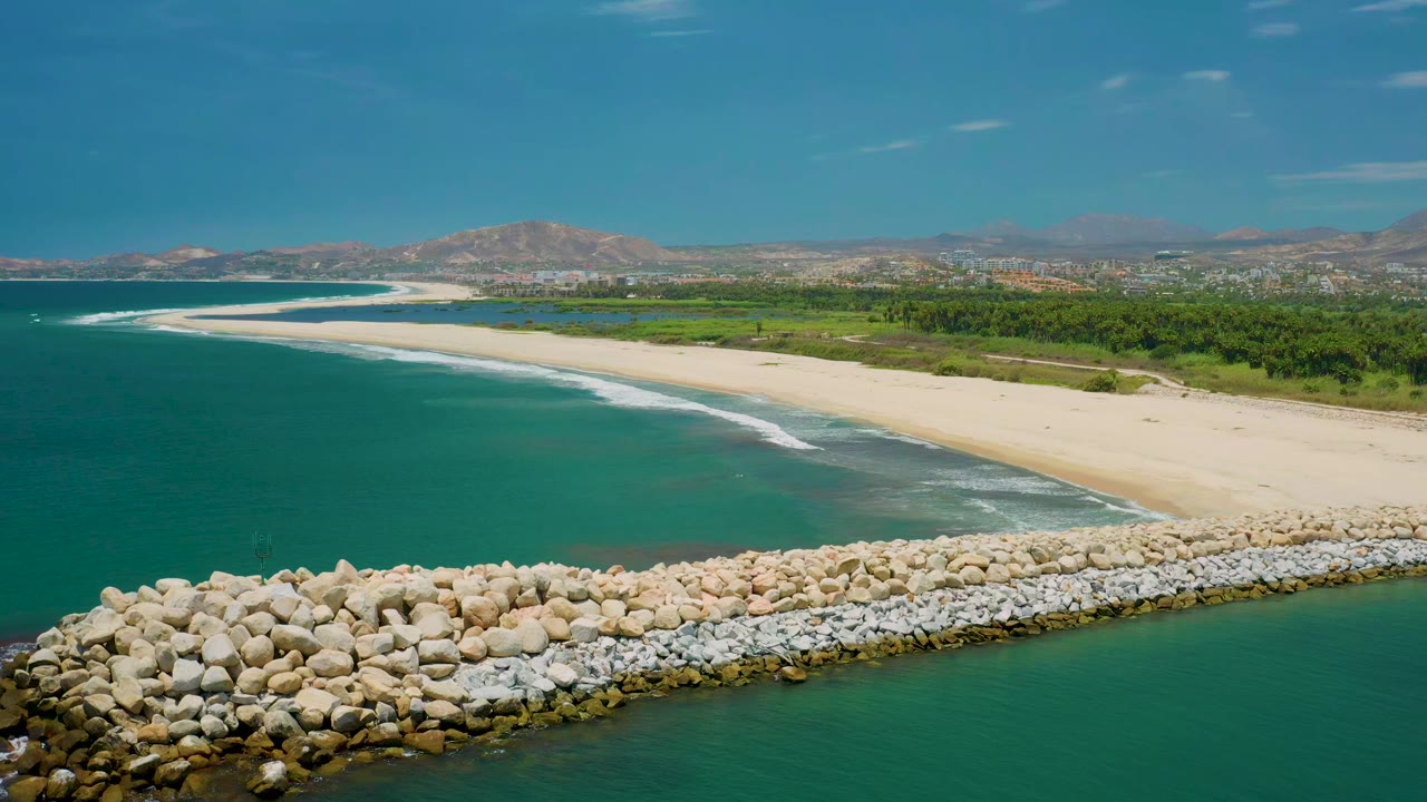 Aerial view of San Jose del Cabo shoreline with the breakwater filling the lower edge, white wave lines along the beach, green inland strip, distant mountains, and no visible people.
