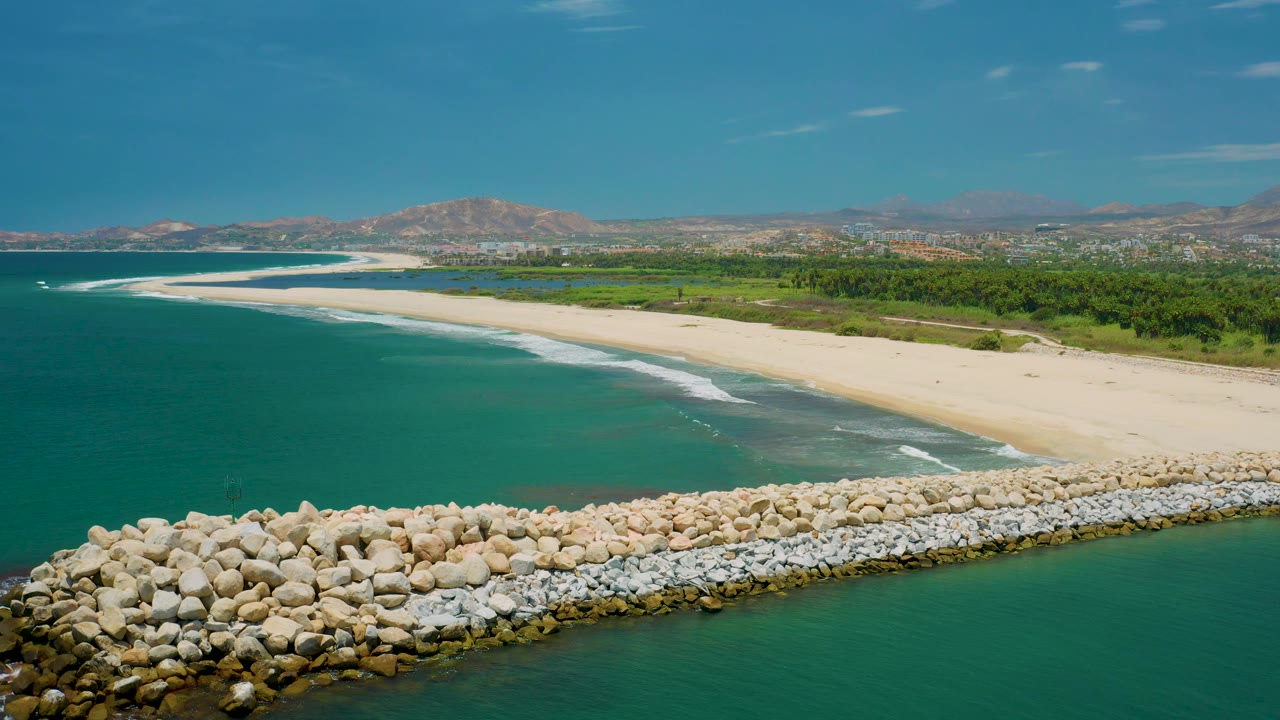 Aerial San Jose del Cabo beach view where the rock breakwater runs across the lower frame, turquoise water meets small white surf, and the beach leads back to greenery and hills with no visible people.