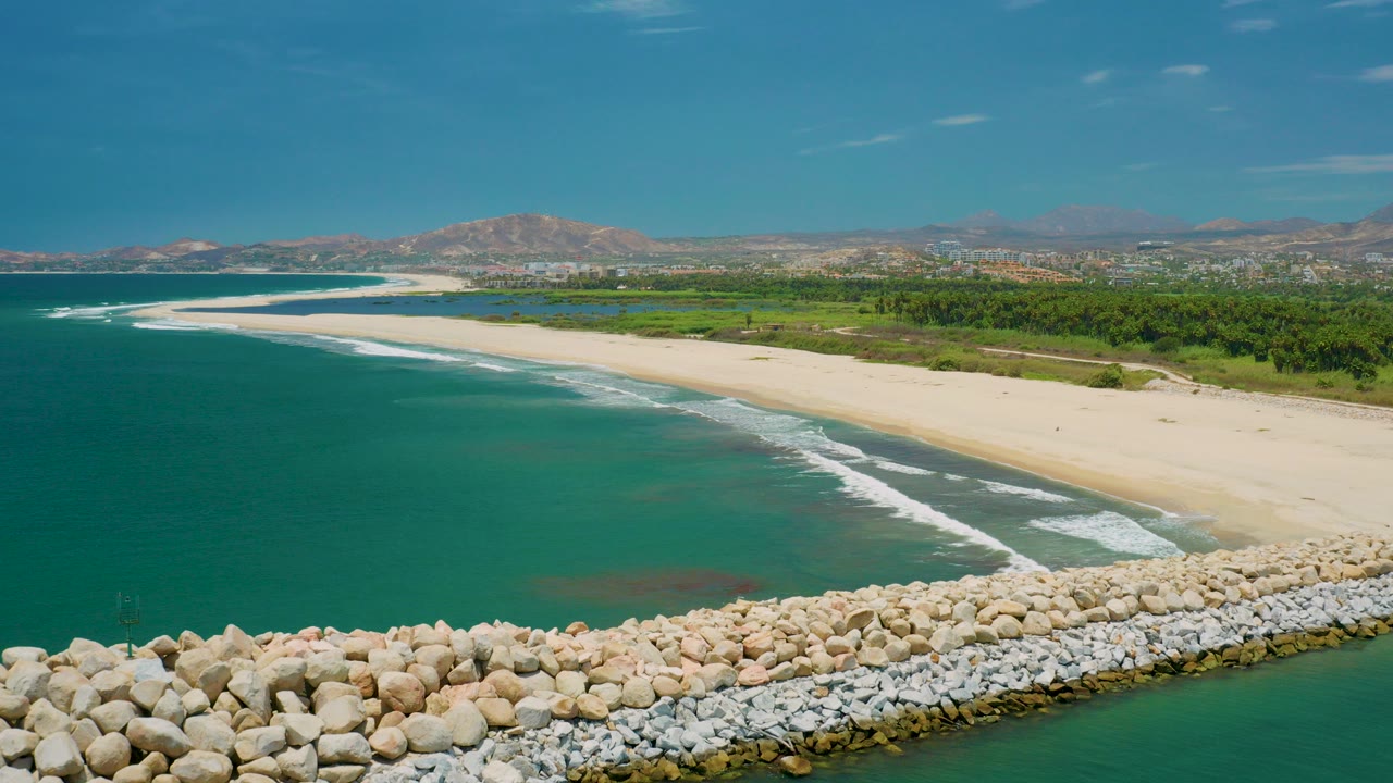 Aerial view in San Jose Del Cabo, Mexico with a rocky foreground, turquoise ocean, a pale sandy beach, green inland trees, distant buildings, and mountains, with no visible people.