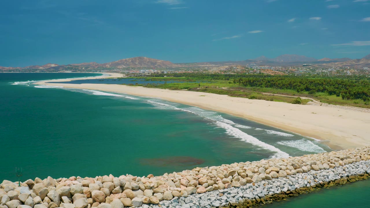 Elevated San Jose Del Cabo, Mexico coast view with darker blue ocean at left, pale sand at center, small surf lines, rock breakwater at the lower edge, inland trees, and mountains in the distance, with no visible people.