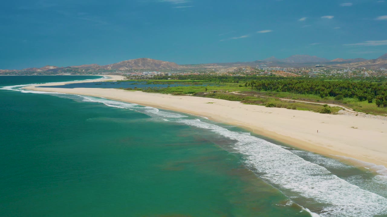 Aerial frame over San Jose Del Cabo, Mexico with turquoise water in the foreground, a white surf line on pale sand, green inland vegetation, distant buildings, hills on the horizon, and no visible people.
