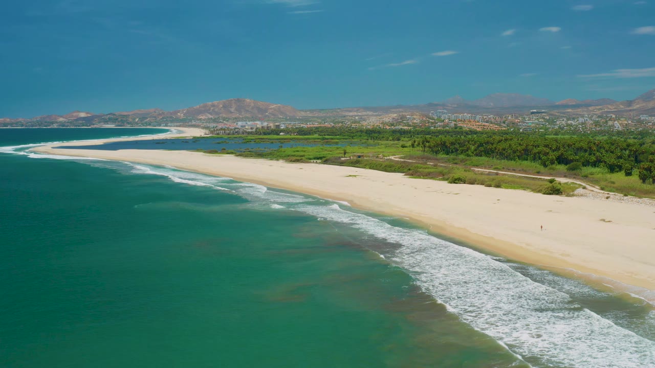 Aerial view over San Jose Del Cabo, Mexico with white foam curving along the beach, darker turquoise water offshore, green land behind the sand, a distant built-up horizon, mountains, and no visible people.