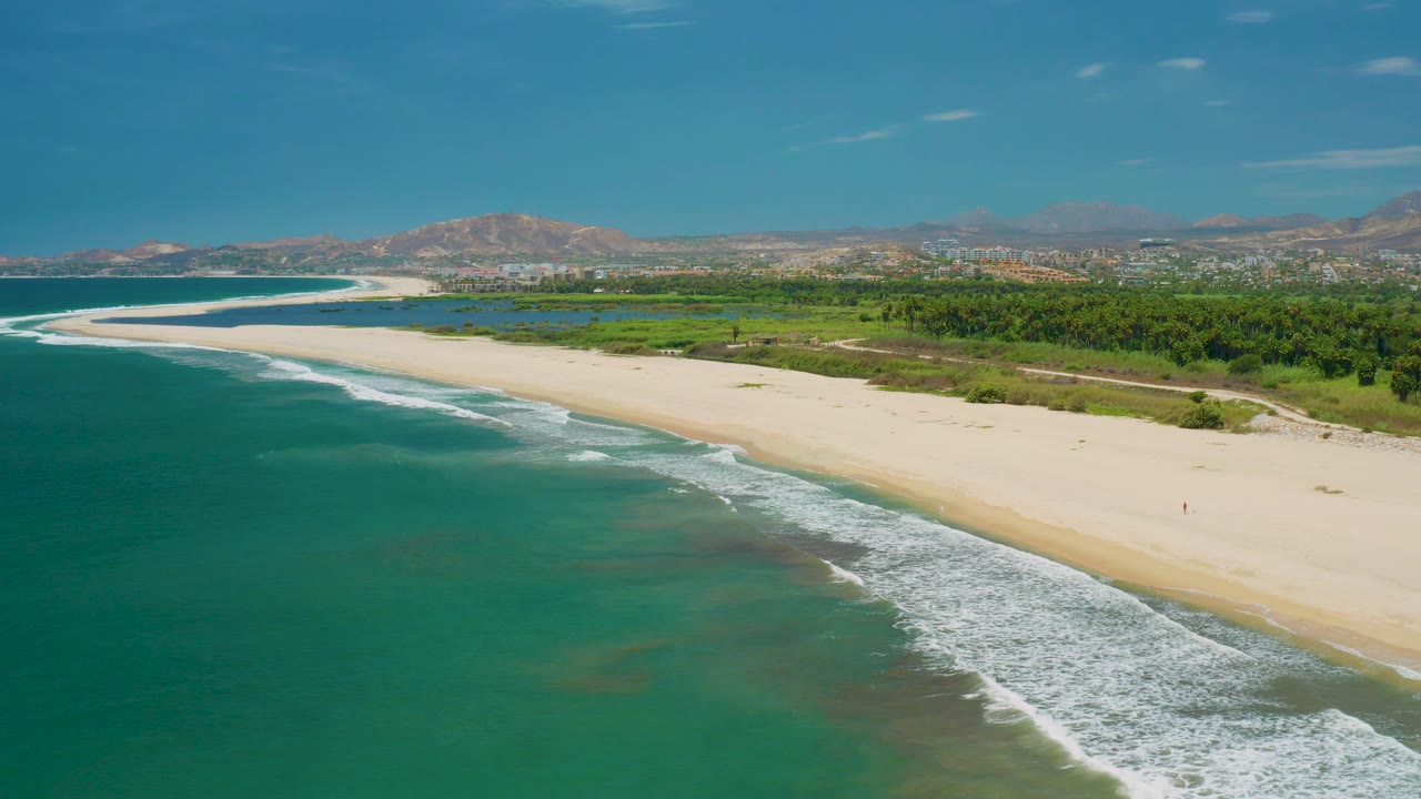 Aerial shoreline scene with ocean foreground, rolling surf, empty sand, trees, and hills.