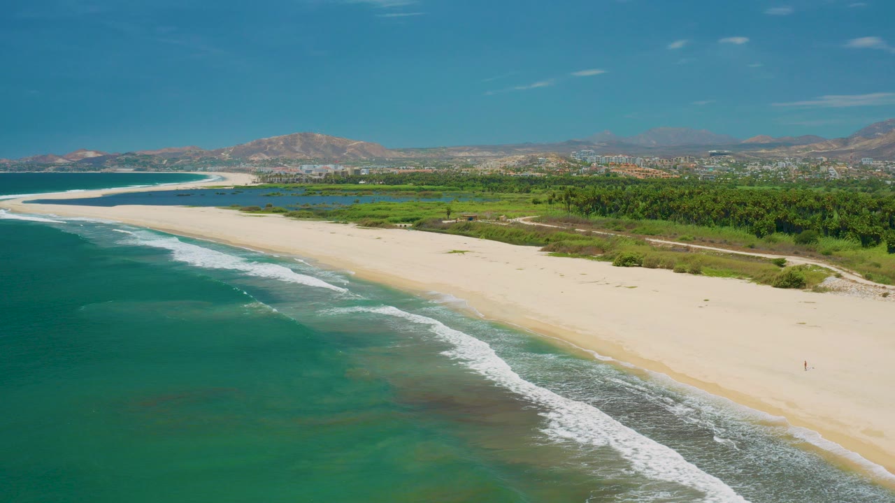 Aerial view of turquoise surf, pale sand beach, green coast, and distant mountains.