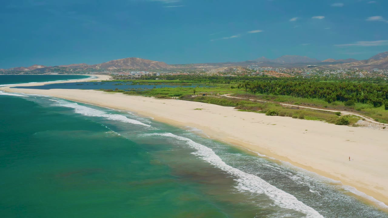 Aerial view of San Jose Del Cabo beach with diagonal white surf, open sand, green coastal vegetation, distant buildings near the horizon, mountains, and no clearly visible people.
