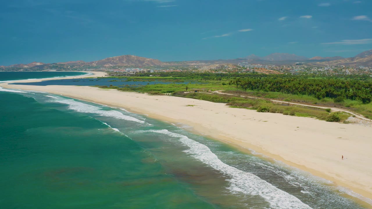 High coastal frame over San Jose Del Cabo showing deeper teal water in the foreground, wave bands on the beach, a green inland band, distant city structures, mountains, and no clearly visible people.