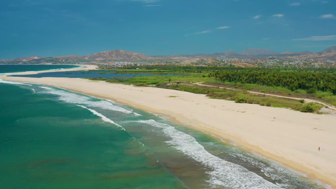 San Jose Del Cabo aerial frame with foamy surf in the lower left, broad white sand across the center, green coastal land and a light path inland, distant buildings, mountains, and no clearly visible people.
