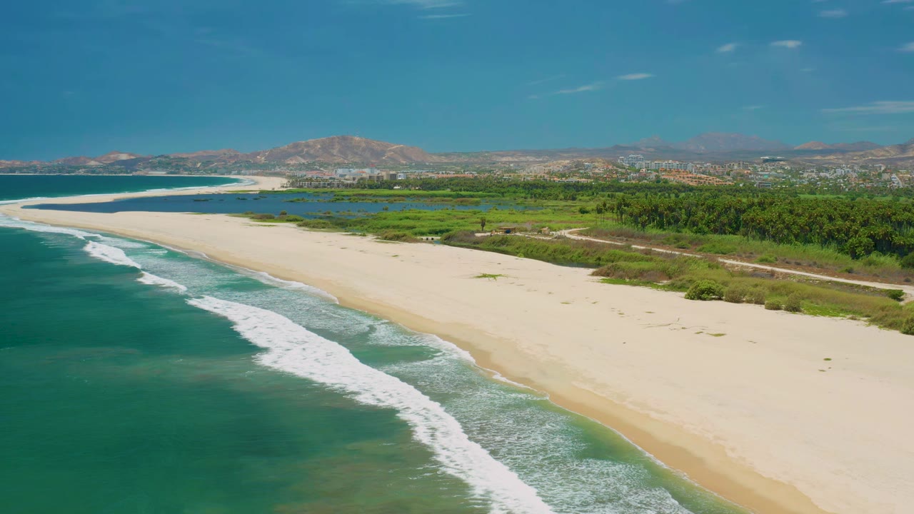 Aerial view of San Jose del Cabo shoreline as the surf line sits farther down the beach, with empty sand, green wetland areas, distant buildings, hills, and no visible people.