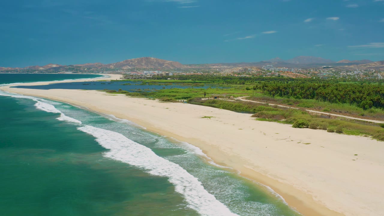 San Jose del Cabo beach seen from above with a long empty sandbar, white waves curling in from the left, green vegetation beyond the beach, distant hills, and no visible people.