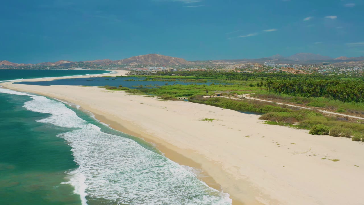 Wide beach and curling surf with green inland vegetation and hills beyond.