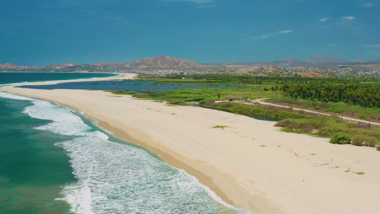 Aerial San Jose del Cabo shoreline with blue-green water and white breakers on the left, empty sand across the center, green brush and hills beyond, and no visible people.