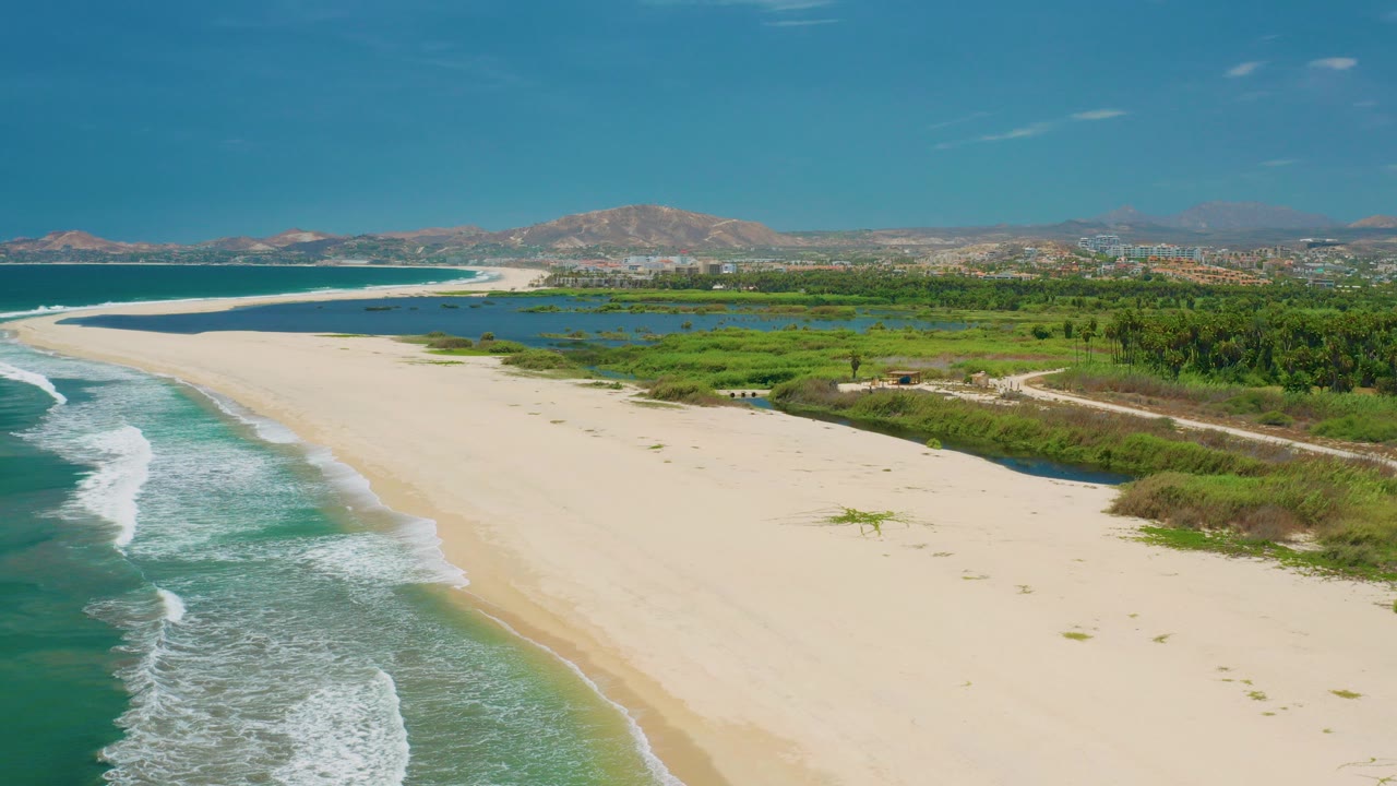 Aerial view of empty beach, surf, lagoon, and distant hills.