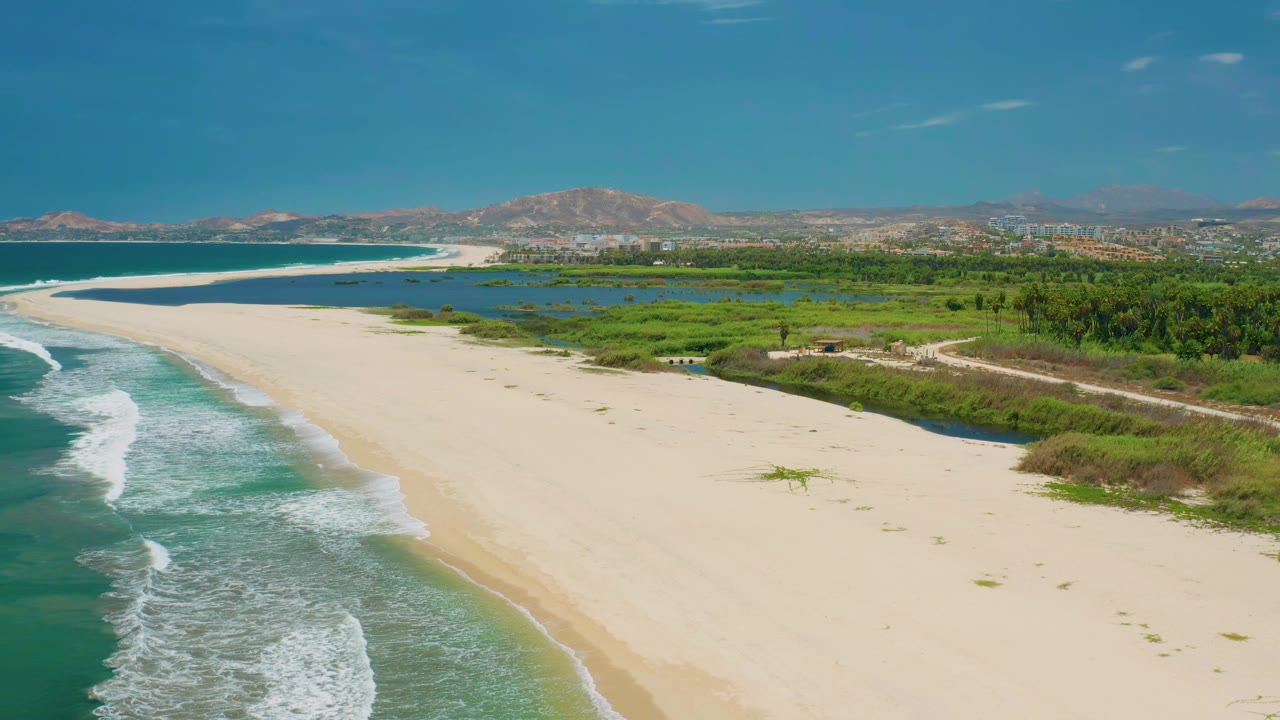 San Jose Del Cabo aerial view with white surf advancing along the left edge, broad pale beach in the center, inland green wetlands, distant mountains, and no visible people.