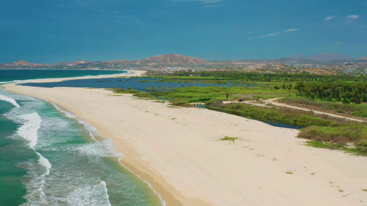 Aerial view over San Jose Del Cabo coastline where the empty beach curves beside foamy surf, with lagoon water and low vegetation leading toward hills in the background and no visible people.