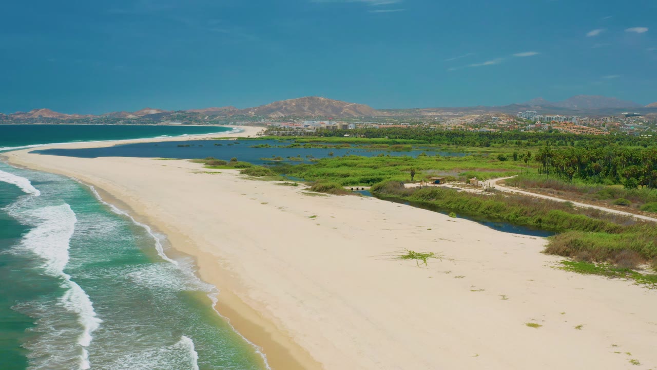 San Jose Del Cabo beach aerial with waves breaking near the lower left, an empty expanse of sand across the middle, lagoon water behind the dunes, distant hills, and no visible people.