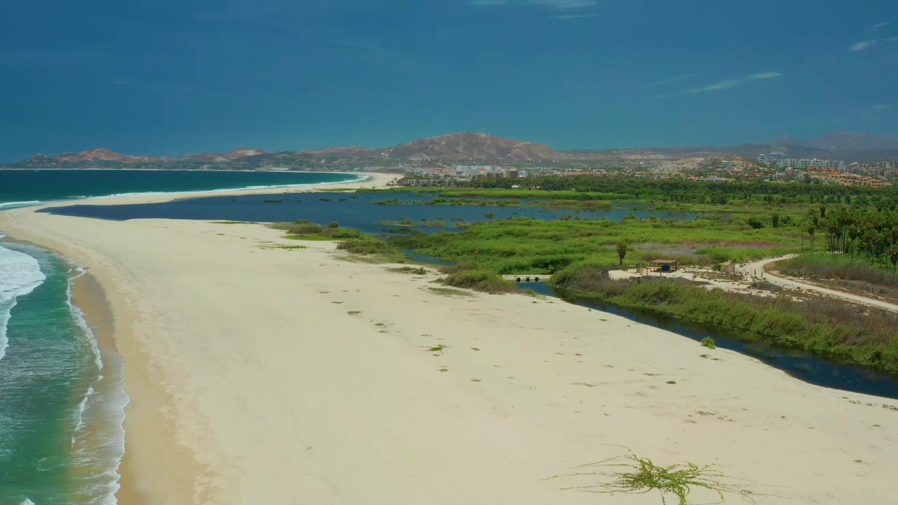Wide aerial shoreline with pale sand, surf, lagoon, and green vegetation.