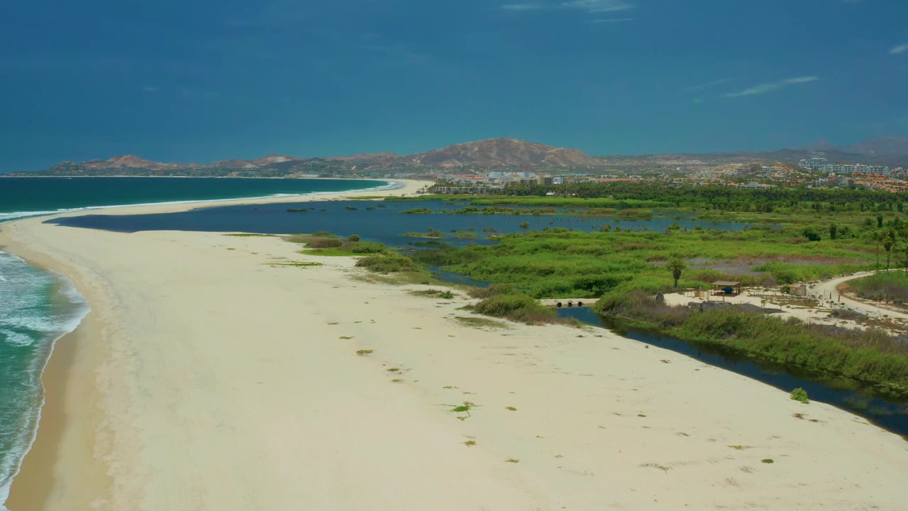 Wide aerial frame in San Jose Del Cabo, Mexico, with ocean surf along the left edge, a broad sandbar through the center, marsh water and vegetation to the right, and no visible people.