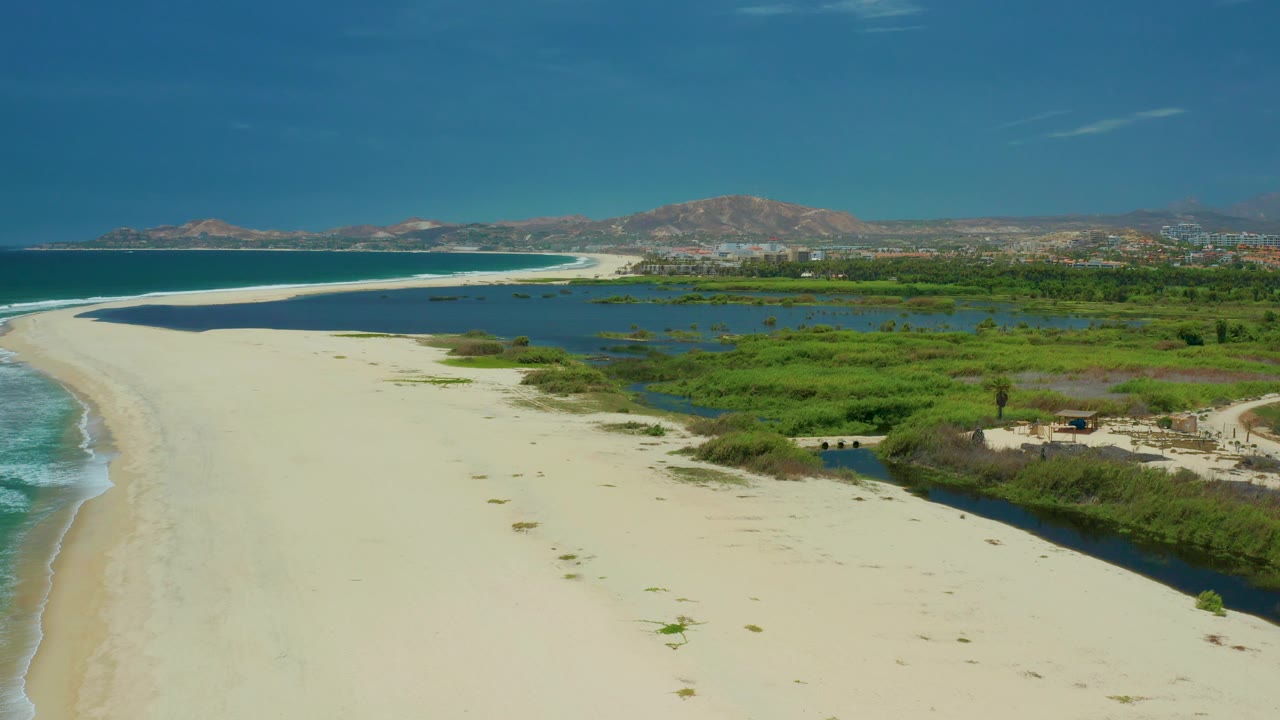 Aerial view in San Jose Del Cabo, Mexico, with the beach widening in the foreground, marsh water and reeds to the right, a small dark inlet near the lower right, and no visible people.