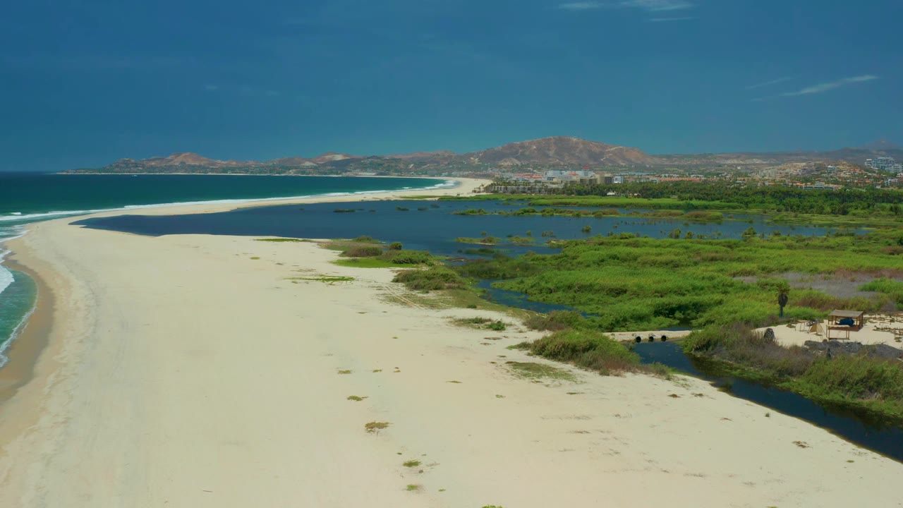 Aerial view in San Jose Del Cabo with the sand curve more centered, ocean on the left, marsh water to the right, mountains beyond, and no visible people.