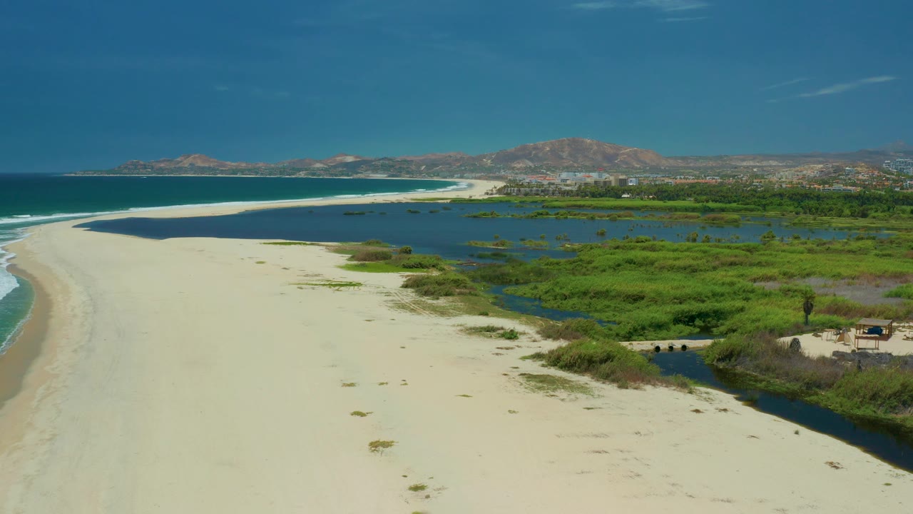 San Jose Del Cabo aerial frame showing a broad pale sand beach, calm blue lagoon water, green wetland patches, low mountains, and no visible people.