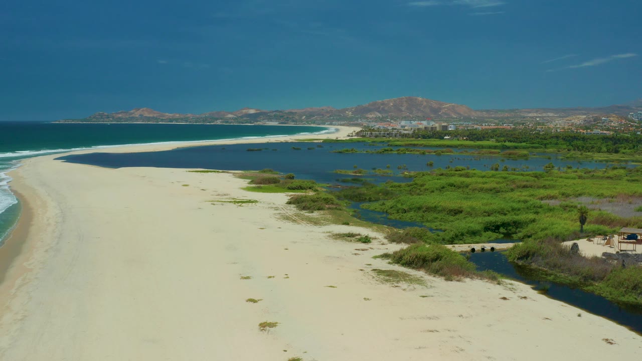 Wide San Jose Del Cabo coastline frame with ocean surf at left, marsh channel and small bridge at lower right, distant mountains, and no visible people.