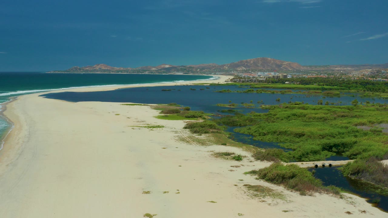 Aerial frame from San Jose del Cabo showing pale sand curving between dark blue ocean and green tidal marsh, with distant hills and no visible people.