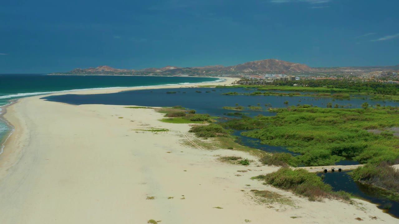 Wide drone view of beach, lagoon water, marsh plants, and mountains.
