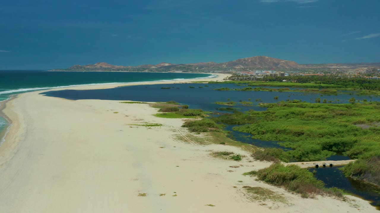 Aerial view over San Jose del Cabo shoreline where light sand separates surf from dark lagoon water and dense green marsh, with no visible people.