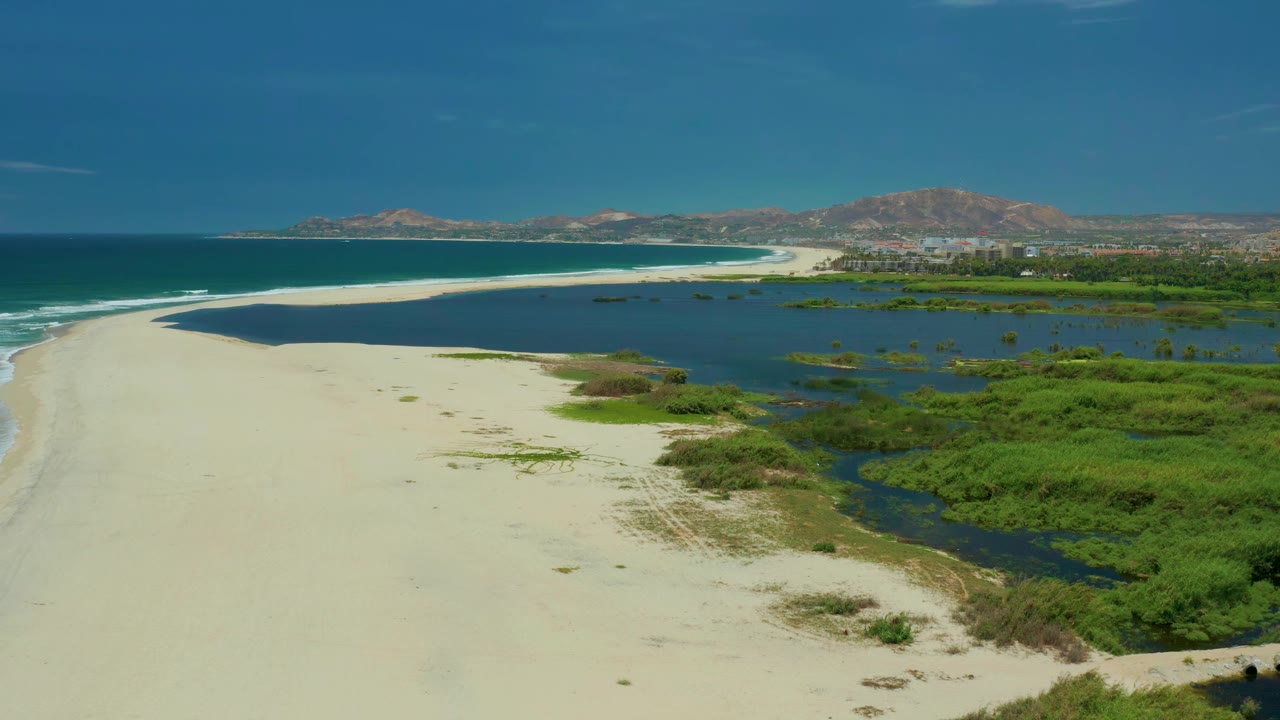 Wide aerial coastline with pale sand, blue ocean, lagoon, marsh vegetation, hills, and distant buildings.