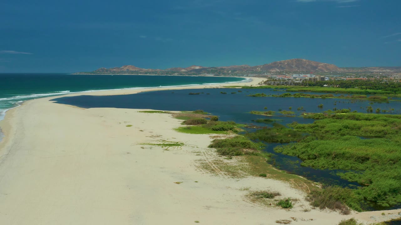At 0:02, San Jose del Cabo appears from above with the beach sweeping toward the horizon, a dark lagoon bordered by green vegetation, ocean waves on the left, and no visible people.
