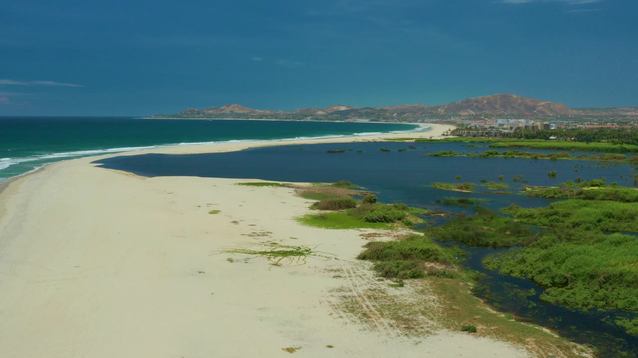 Aerial frame over San Jose Del Cabo showing pale sand in front, dark lagoon water beside green vegetation, ocean surf at left, distant hills and buildings, and no visible people.