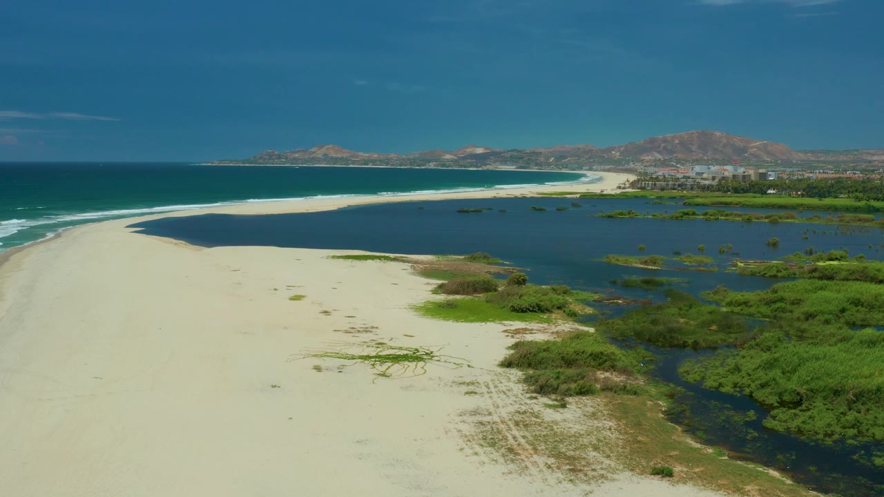Aerial frame over San Jose Del Cabo where bright sand fills more of the lower frame, the lagoon curves beside green patches, the surf line sits left, and no visible people appear.
