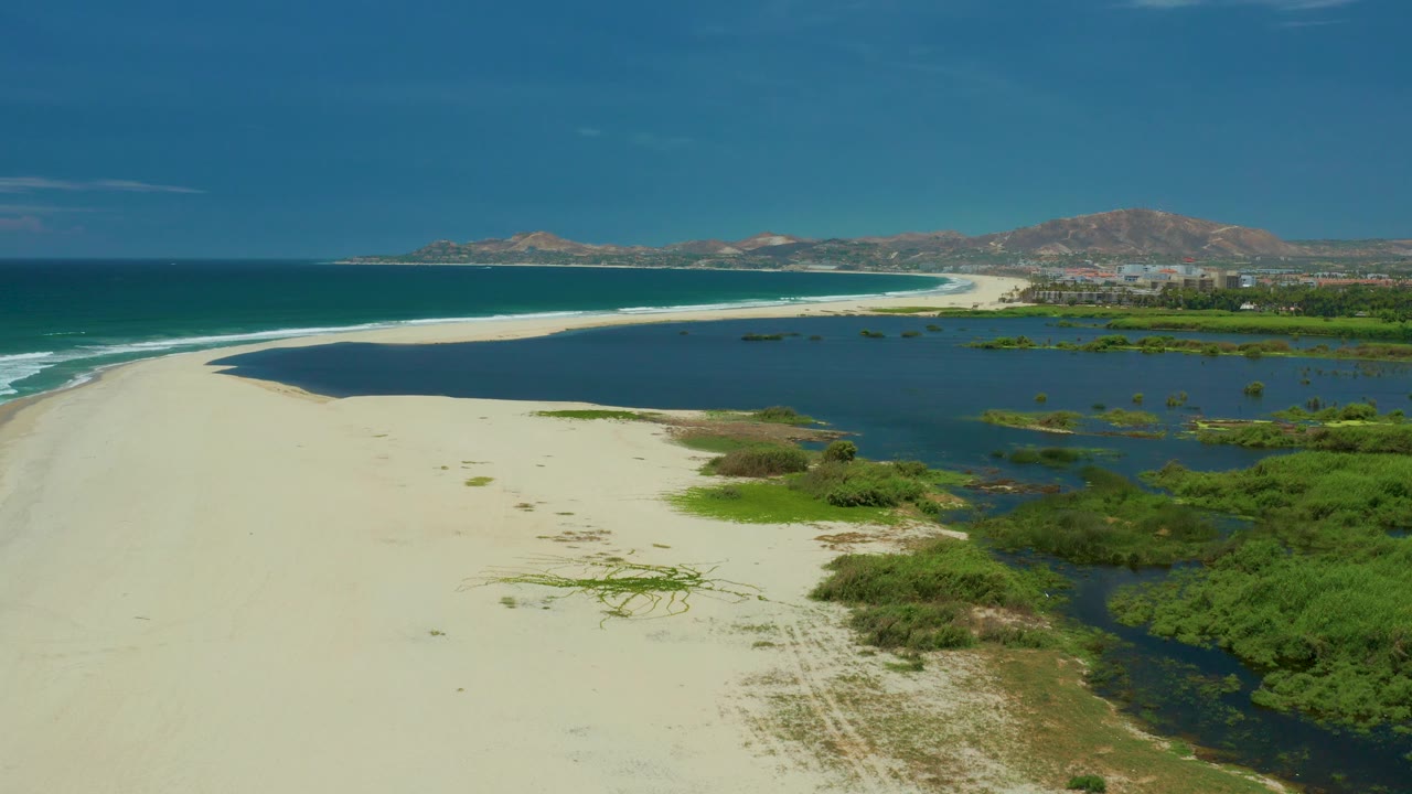 Aerial frame in San Jose Del Cabo with the pale beach sweeping from the lower left, lagoon water and wetland greenery to the right, surf at far left, and no visible people.