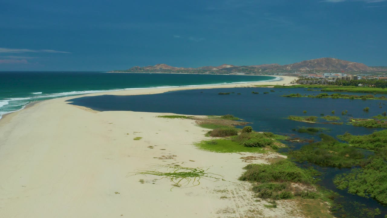 Aerial frame from San Jose Del Cabo showing pale sand between blue ocean surf and dark lagoon water, with green shoreline bushes, distant hills, and no visible people.