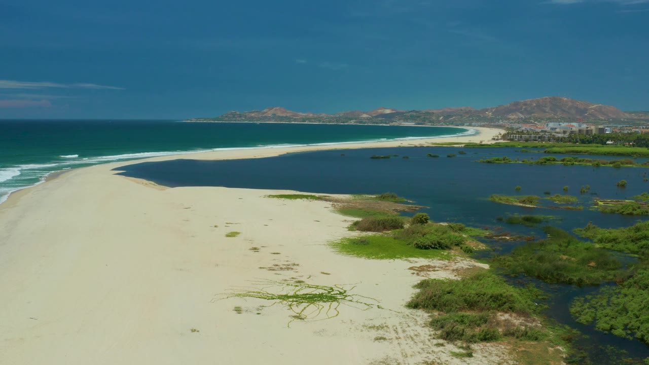 Aerial frame over San Jose Del Cabo beach with empty pale sand in the foreground, blue sea and small white waves to the left, lagoon water to the right, and no visible people.
